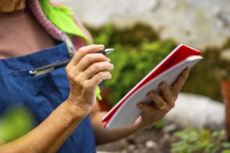 Senior woman in apron and colorful scarf writing in a notebook with pen while observing plants in a