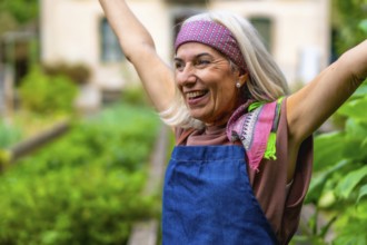 Senior woman in a bandana and apron raises her arms in a sunlit garden, smiling with joy and