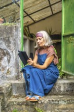 Senior woman in denim overalls and headband sits on rustic outdoor steps in a garden, focused on a