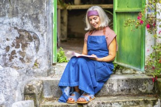 Senior woman in denim apron sits on old stone steps in a sunny garden, peacefully reading a book,