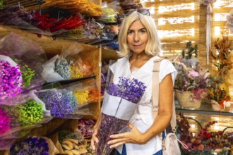 Mature woman with blond hair holds a purple dried flower bouquet inside a colorful market stall
