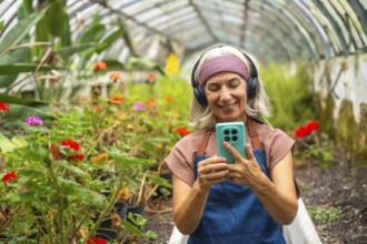 Senior woman wearing headphones and apron, smiling while looking at her smartphone and interacting
