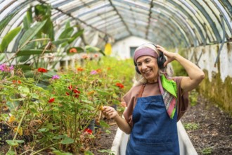 Senior woman smiling, wearing an apron and headphones, happily tending to potted plants and flowers