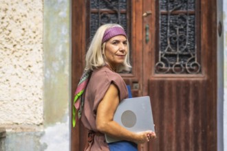 Senior woman with gray hair wearing a headband and casual outfit, carrying a laptop and looking