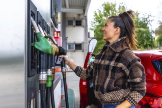 Woman in a casual plaid jacket holding a fuel pump nozzle, refueling her red car at a gas station,