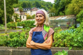 Senior woman wearing an apron and bandana standing with arms crossed in a vibrant community garden,