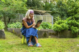 Senior woman sitting on a stool in her green garden, using a digital tablet for managing her plants