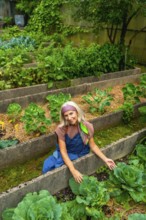 Senior woman in overalls smiling while tending raised bed allotment, harvesting organic vegetables