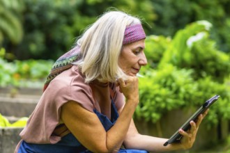 Senior woman with blond hair and a pink headband enjoying leisure time in a lush green garden,