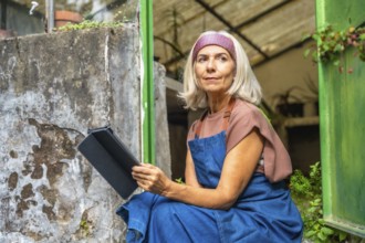 Senior woman in apron sitting by a greenhouse entrance, holding a digital tablet and looking