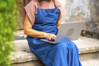 Senior woman in a blue apron working on a laptop outdoors, managing her small business and staying