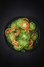 Sliced green tomatoes, on a dark surface, garnished with herbs and salt, top view