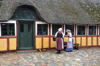 Two women in older farm costumes in front of a half-timbered house with a thatched roof and on