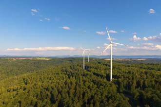 Wind turbines over green forest area, surrounded by beautiful clouds under a blue sky, near
