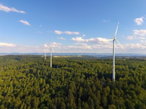 Three wind turbines stand in a green forest landscape under a clear sky, near Schorndorf,