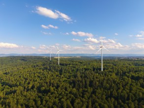 Several wind turbines rise above a lush forest under a sky with light clouds, near Schorndorf,