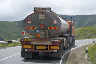 A tank truck drives on a winding road through a mountainous and green landscape under cloudy sky,