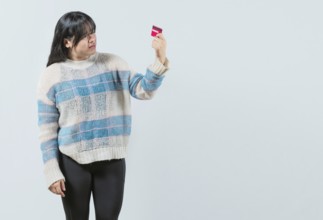 Unsure and confused young woman holding and looking at a credit card isolated. Confused asian girl