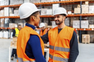 Two diverse warehouse workers wearing safety hard hats and vests are confidently smiling and
