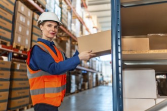 Woman worker wearing safety helmet and reflective vest, putting a cardboard box on a shelf in a
