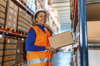 Professional female warehouse worker in hard hat and safety vest holding a carton, smiling