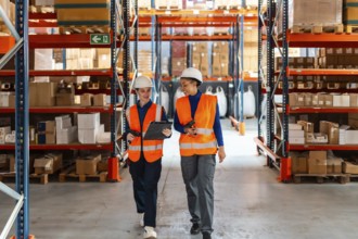 Two diverse female workers wearing safety vests and hard hats, discussing inventory management