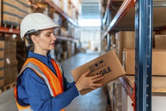 Woman warehouse worker wearing safety helmet and vest, inspecting a cardboard box with fragile