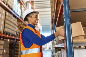 Smiling young woman, wearing a safety helmet and high visibility vest, arranging cardboard boxes on