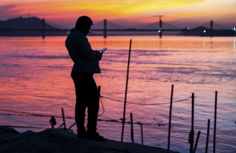 A person browsing his mobile phone by the Brahmaputra riverside, during sunset, on November 4, 2025