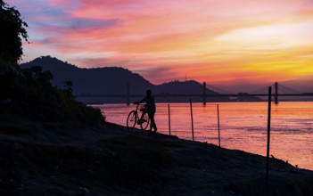 A man pushes his bicycle along the Brahmaputra riverbank during a vibrant sunset, with the