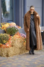 A woman stands next to the autumn installation, wearing a brown coat and black dress. She poses