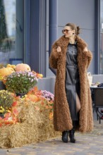 A woman stands confidently in a warm, fluffy coat at an outdoor market location surrounded by