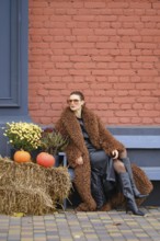A woman sits on a bench dressed in warm fur coat. She poses near colorful pumpkins and vibrant