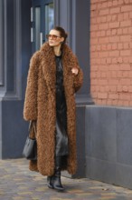 A fashionable woman strolls along a city sidewalk wearing a warm brown fur coat and stylish black