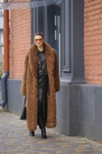 A fashionable woman strolls along a city sidewalk, wearing a cozy brown fur coat. She pairs it with