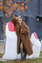 A fashionable woman in a cozy brown coat stands near large white sculptures resembling fingers with