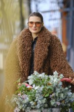 A woman in oversized sunglasses and a fluffy brown coat strolls through a vibrant urban area. She