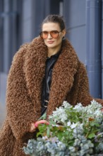 A beautiful woman stands outdoors in a city, wearing a large brown fluffy coat and orange