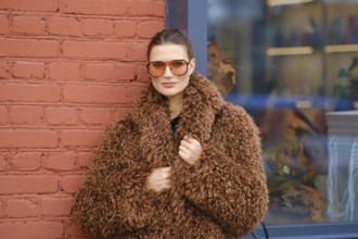 A woman poses confidently in a warm, brown, fluffy coat and oversized sunglasses. She stands