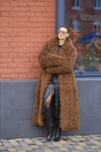 A fashion model stands with crossed arms beside a red brick wall. She wears a fluffy brown coat, a