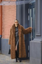 A fashionable woman in a long brown fur coat and sunglasses walks out of a colorful urban office