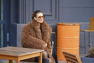 A fashionable woman sits outside a cafe, wearing a large brown fur coat and stylish sunglasses. She
