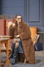 A stylish woman is seated at a wooden table outside a cafe, wearing a fluffy brown coat and
