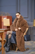 A stylish woman sits at an outdoor cafe during autumn, wearing a fuzzy brown coat and black
