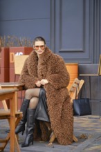 A woman sits comfortably at a wooden cafe table, wearing a brown fluffy coat and knee-high black
