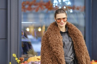 A woman wearing a brown furry coat and sunglasses stands outside a cozy cafe filled with autumn