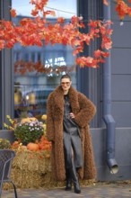 A stylish model stands near a store with bright autumn leaves overhead. She wears a brown fur coat