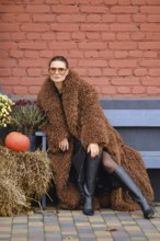 A woman wearing a brown fuzzy coat and knee-high black boots poses confidently outdoors. She is