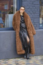 A fashionable woman stands against a brick wall, wearing a warm, fluffy coat over a sleek black