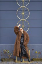 A woman stands confidently near a striking bike wheel art piece. She wears a fluffy brown coat and
